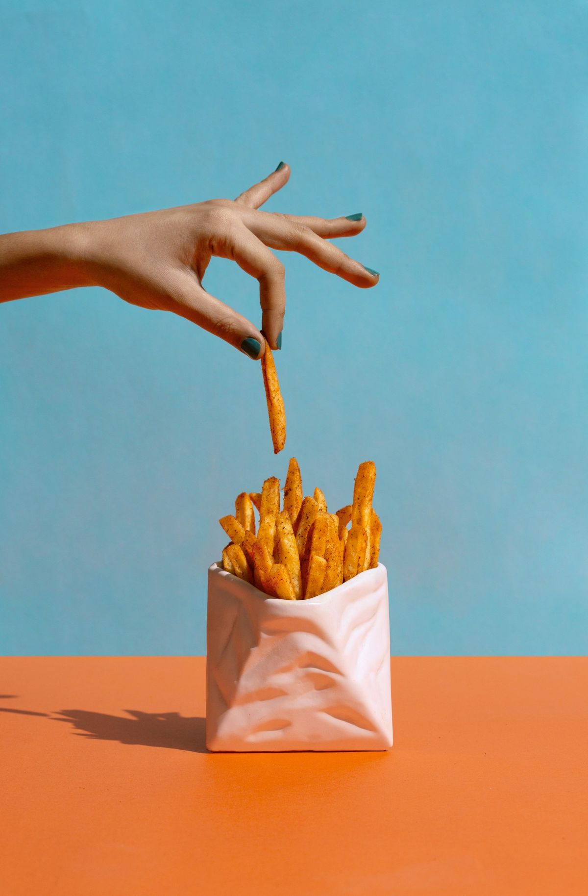 A hand picking a fry from a white paper cone, on a blue and orange backdrop